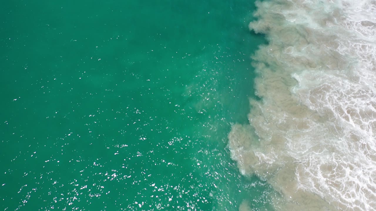 Overhead view of turquoise-green water in Los Cabos, Mexico, with bright surface sparkle and white surf entering from the right, no visible people.