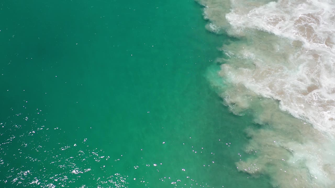 Overhead view of turquoise water in Los Cabos, Mexico, with white surf foam pushing in from the upper right and no visible people.