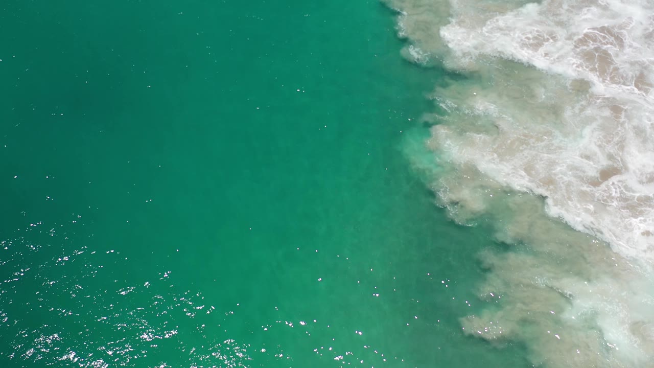 Overhead Los Cabos water frame with green-blue ocean, scattered sunlight reflections near the lower left, and foamy surf along the right side, with no visible people.