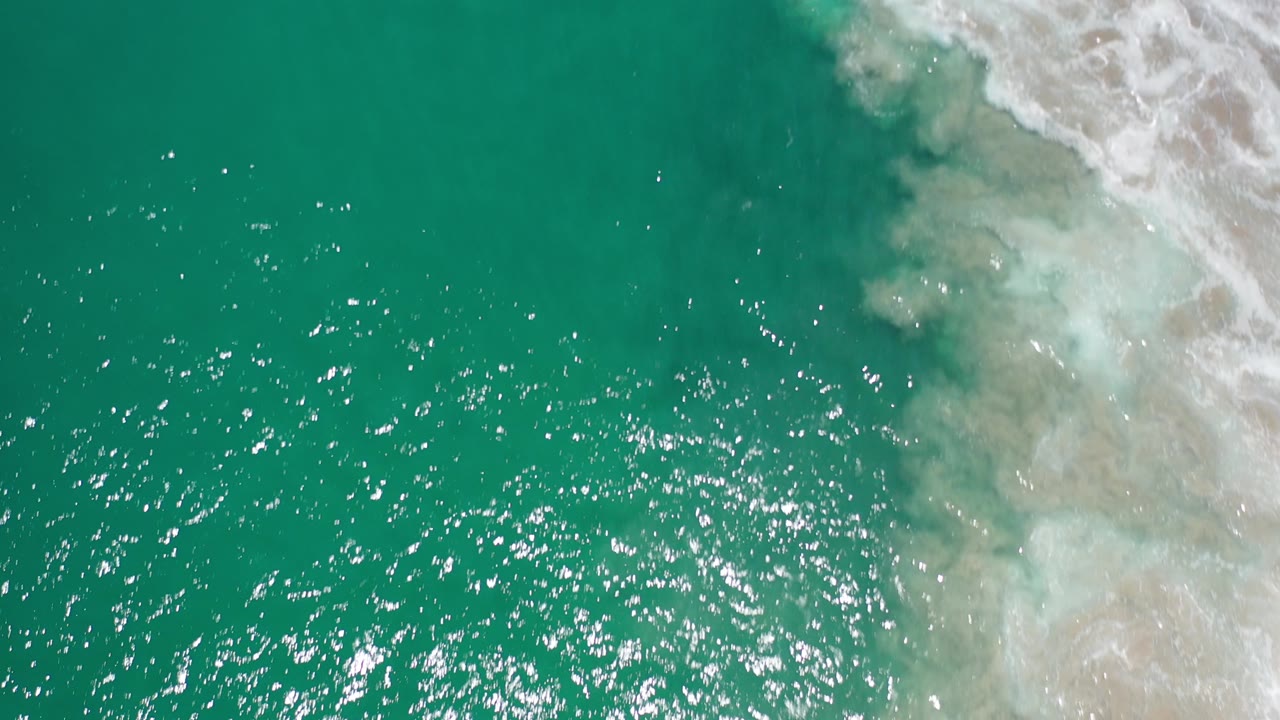 Overhead view in Los Cabos, Mexico of green-blue water with bright sun flecks and pale foamy surf meeting sand on the right, with no visible people.