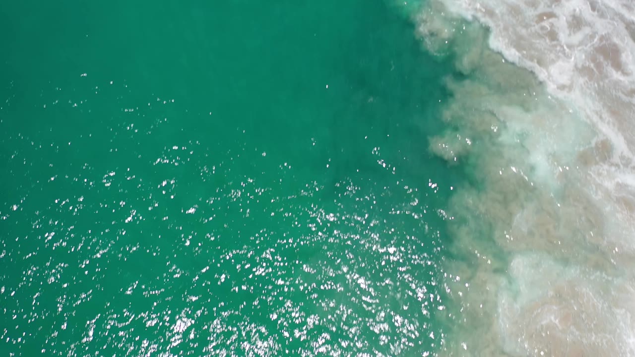 Aerial Los Cabos shoreline frame with rippled green water across the left and center, foamy water over tan sand on the right, and no visible people.