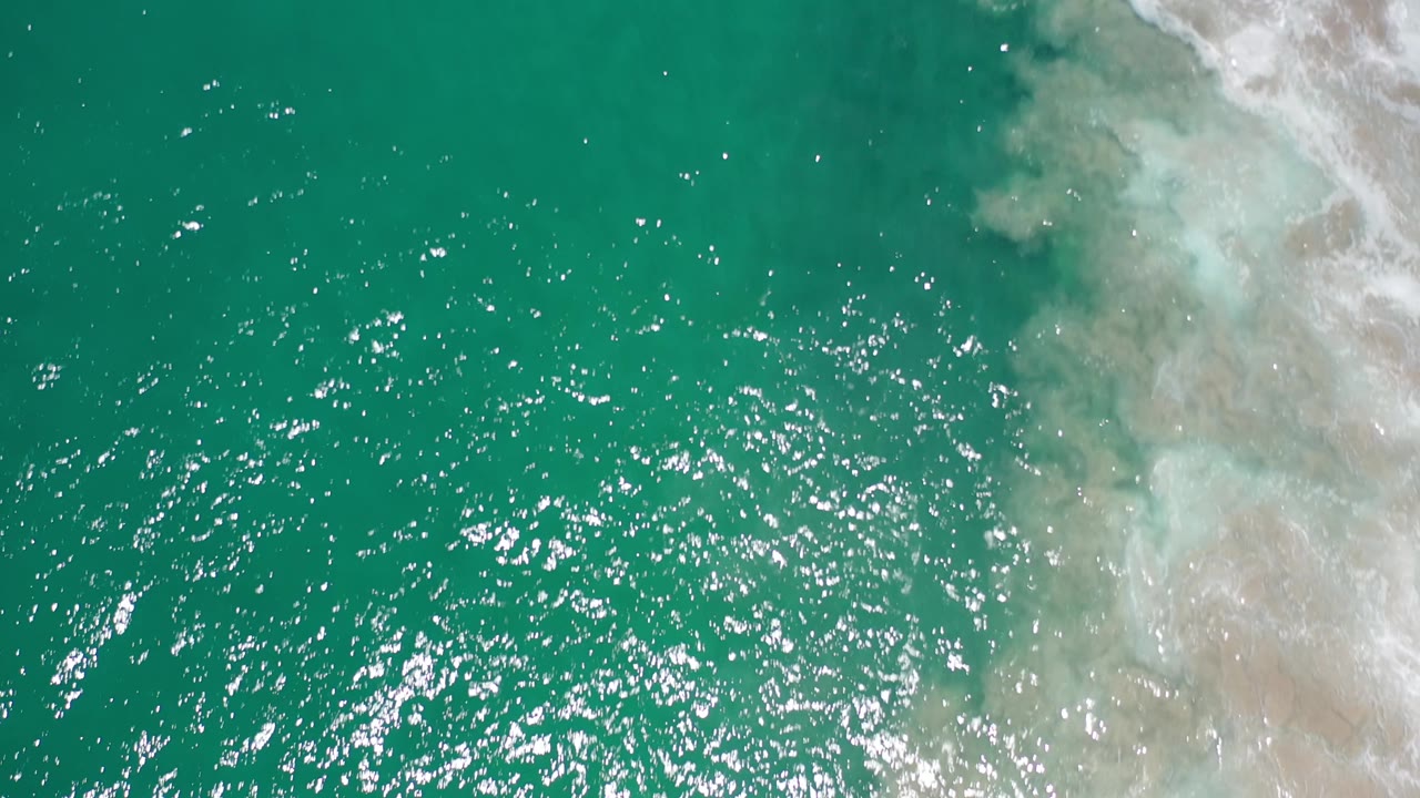 Sunlit green sea surface with white foam over sand on the right.