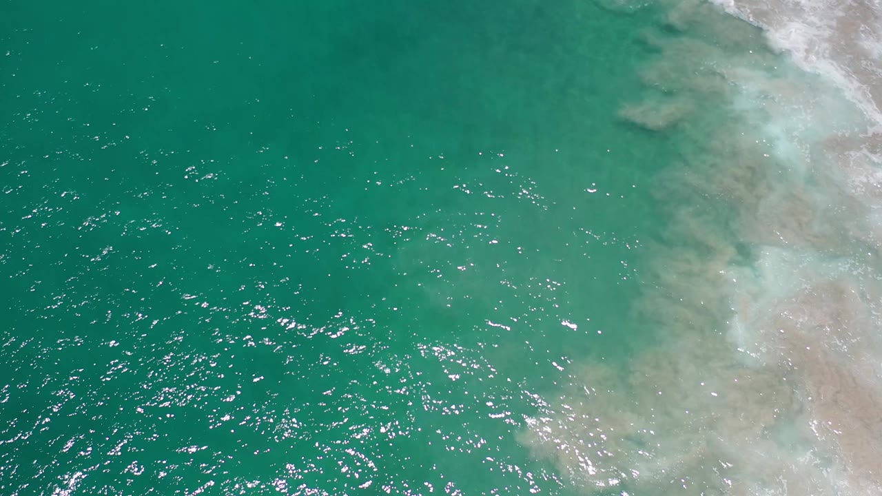 Aerial view in Los Cabos, Mexico, showing green sea surface across most of the frame, scattered white highlights, pale foamy water to the right, and no visible people.
