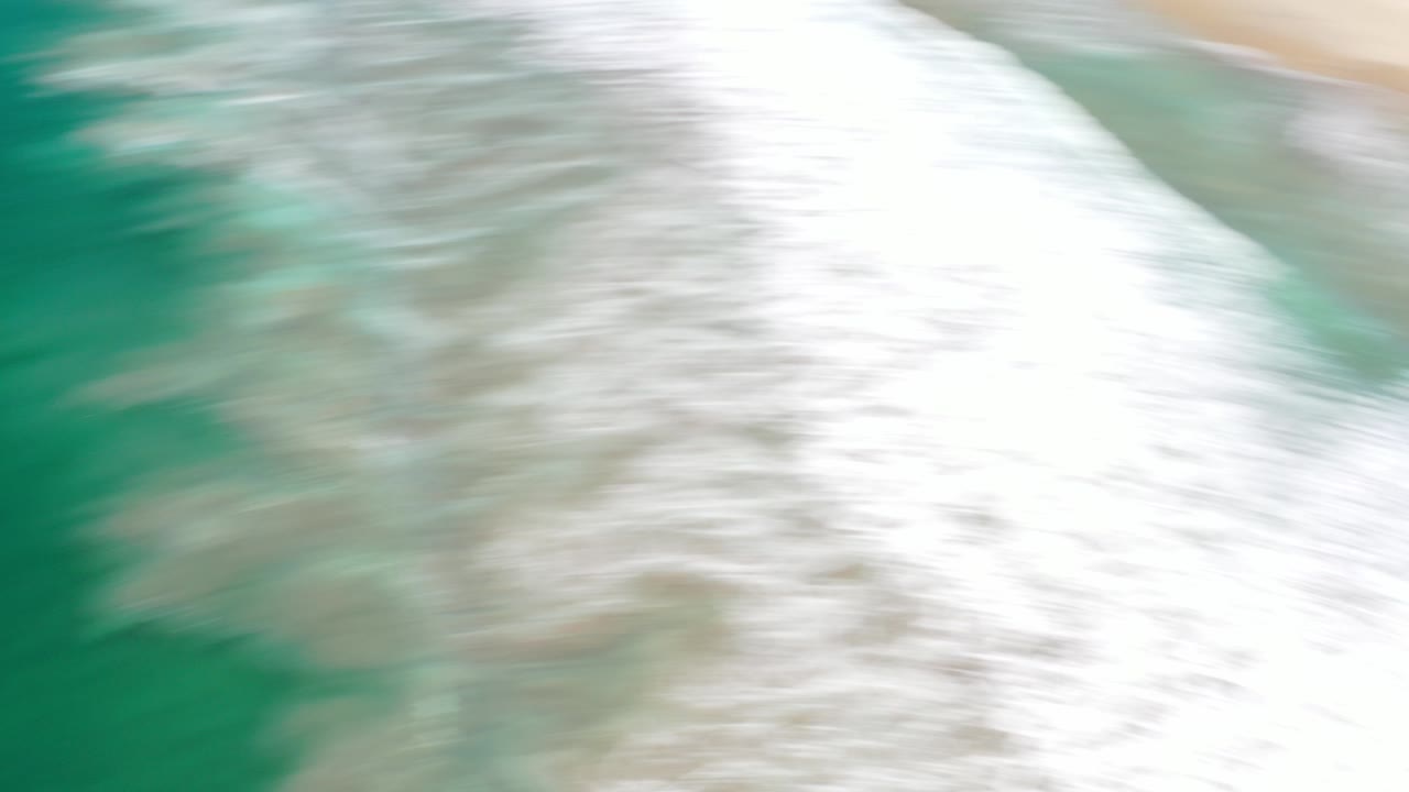 Overhead view of Los Cabos surf blurring across turquoise water beside pale sand, with no visible people.