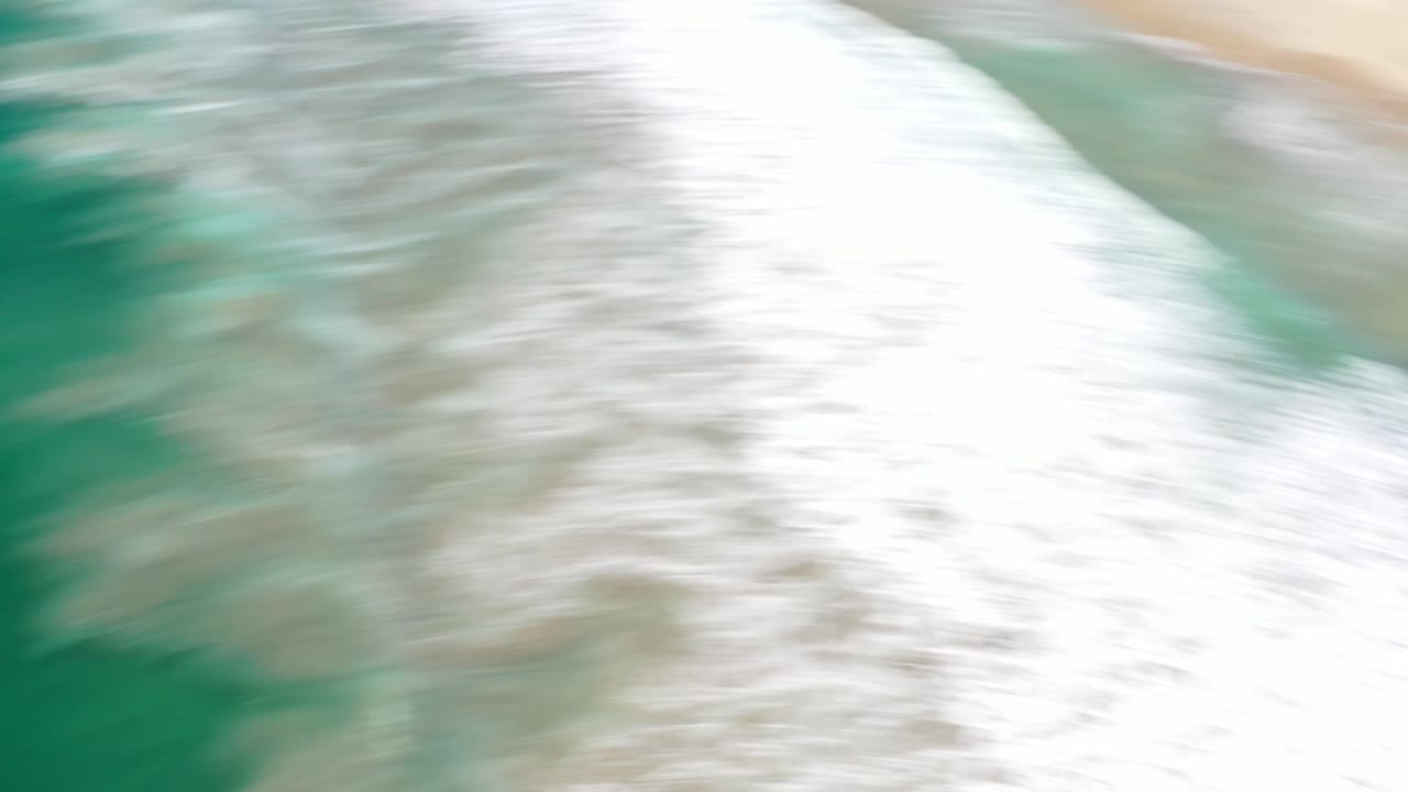 A bright overhead Los Cabos beach frame shows a broad white wave edge pushing toward smooth pale sand, with no visible people.