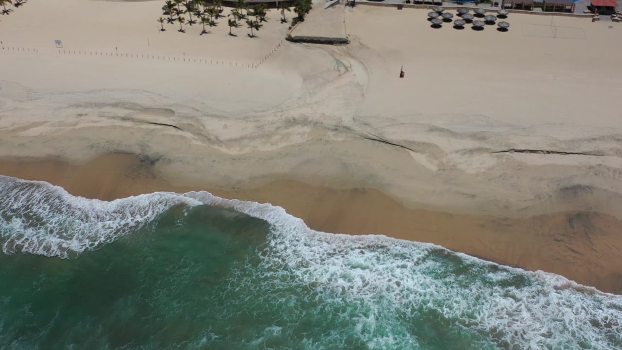 Overhead Los Cabos beach view with green water, white surf, pale sand, palm trees, umbrellas, and resort pools along the top edge, with no visible people.