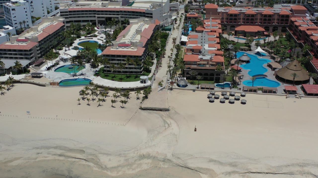 High aerial frame over Los Cabos, Mexico showing pale beach in the foreground, resort pools and red-roofed buildings behind it, with no clearly recognizable people.