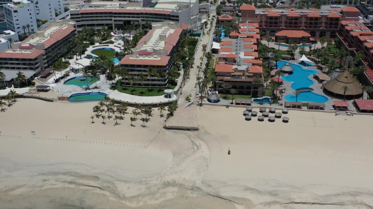 Aerial Los Cabos, Mexico view with a broad pale beach below, a central walkway to the shore, blue pools, red roofs, palms, and tiny distant figures on the sand.