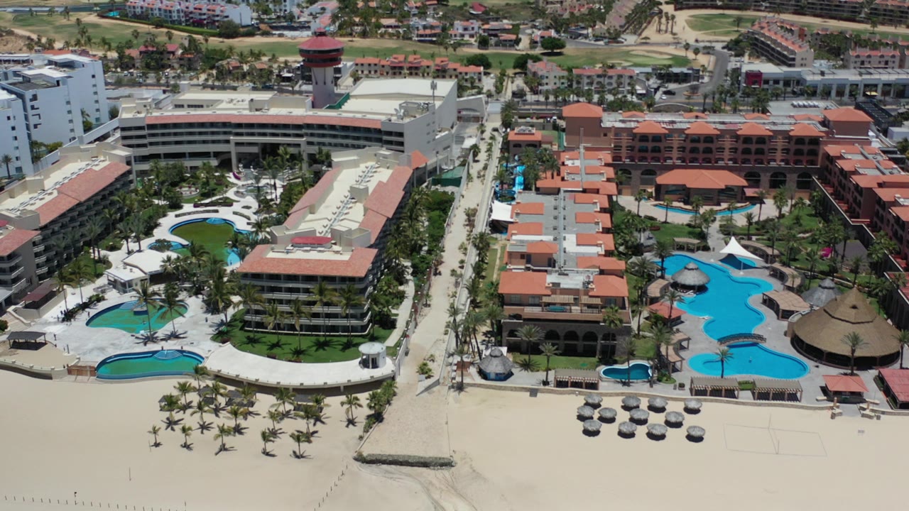 Aerial view of a hotel resort with pools, palms, sand, and red-tile roofs.