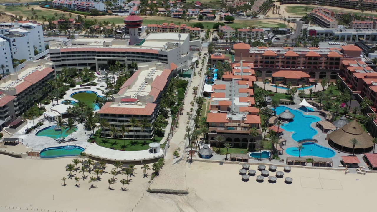 Second Los Cabos aerial frame showing the resort pools slightly farther across the property, with white hotel blocks, palms, sand, and no visible people.