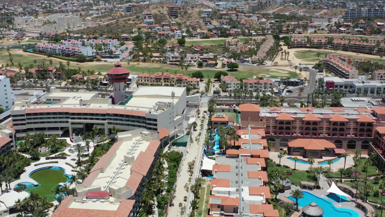 Second aerial frame in Los Cabos with the resort complex lower in frame, curved roads, green fairways, hillside homes, unfinished-looking buildings, and no visible people.
