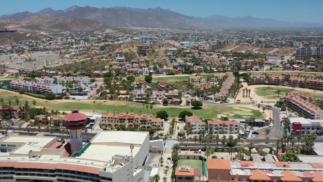 Los Cabos aerial view one second in, showing the red-topped tower near the lower edge, apartment-style buildings, a road grid, green fairways, dry hills, mountains, and no visible people.