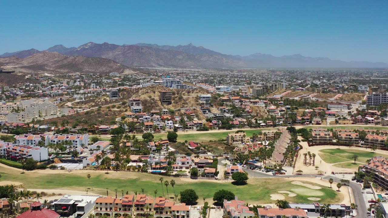 Wide aerial view of neighborhoods, golf greens, roads, and mountains under blue sky.