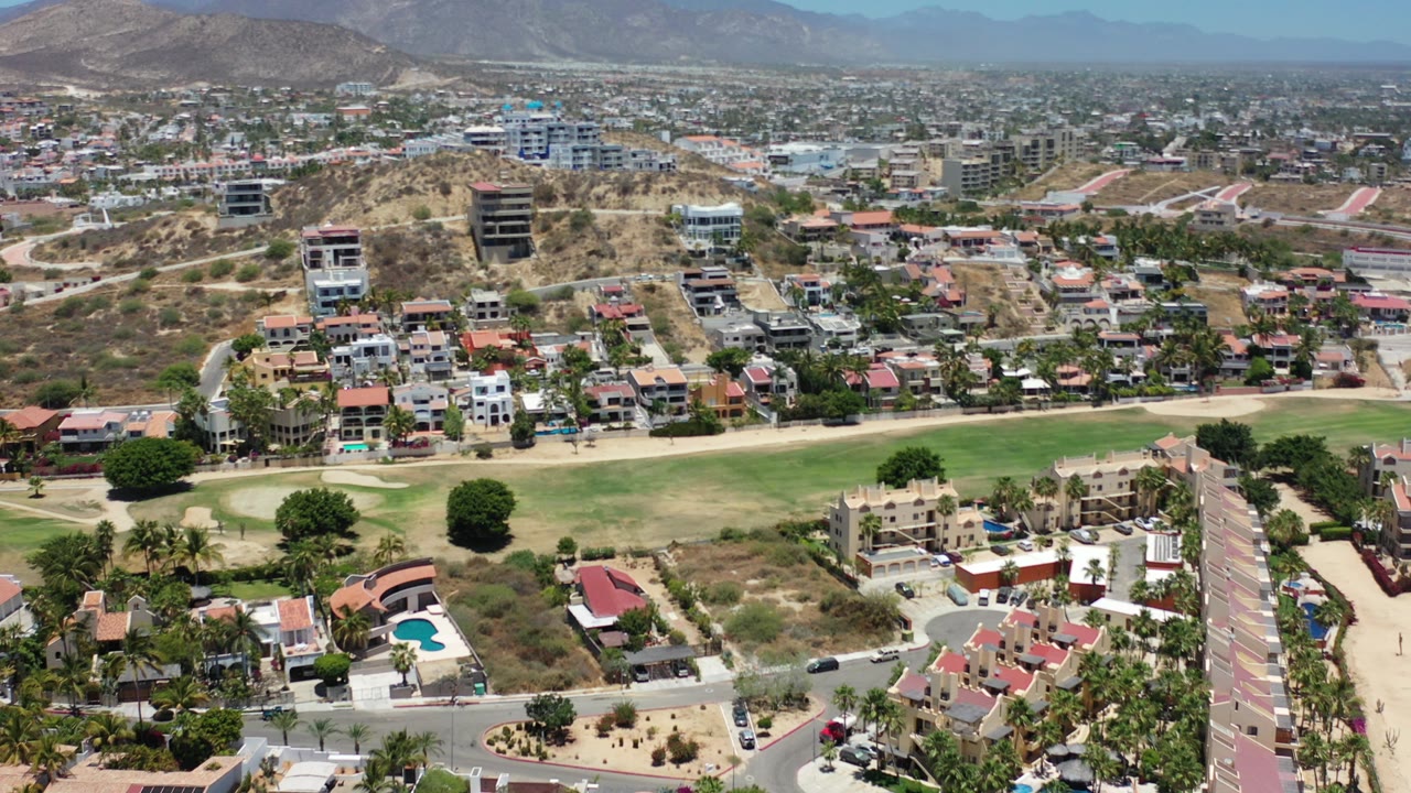 High aerial view over a Los Cabos residential area with red-roofed homes, a green golf fairway, dry hills, distant mountains, and no visible people.