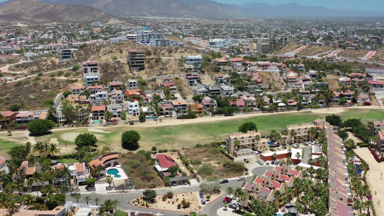 Aerial frame in Los Cabos looking across clustered houses, a pale road, a green fairway, hillside buildings, and hazy mountains with no visible people.