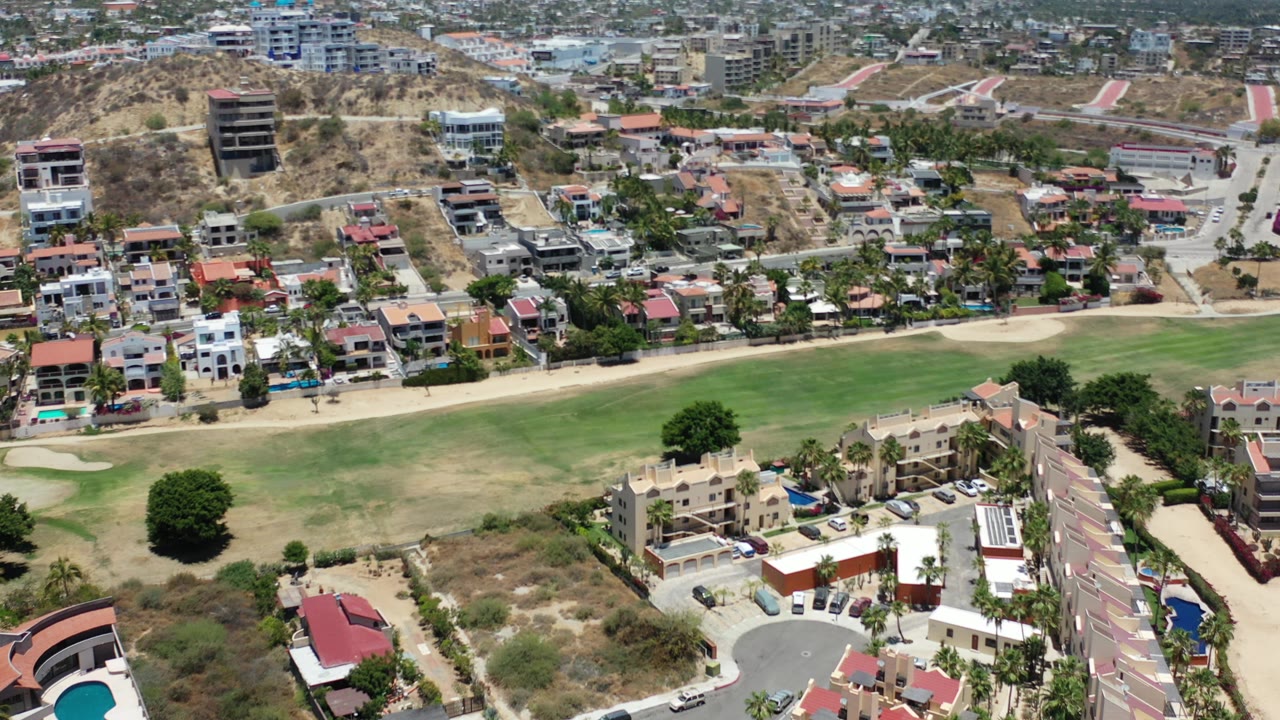 Opening aerial frame over Los Cabos, Mexico, showing hillside residences, paved roads, green open ground, dry slopes, scattered trees, pools, and no visible people.