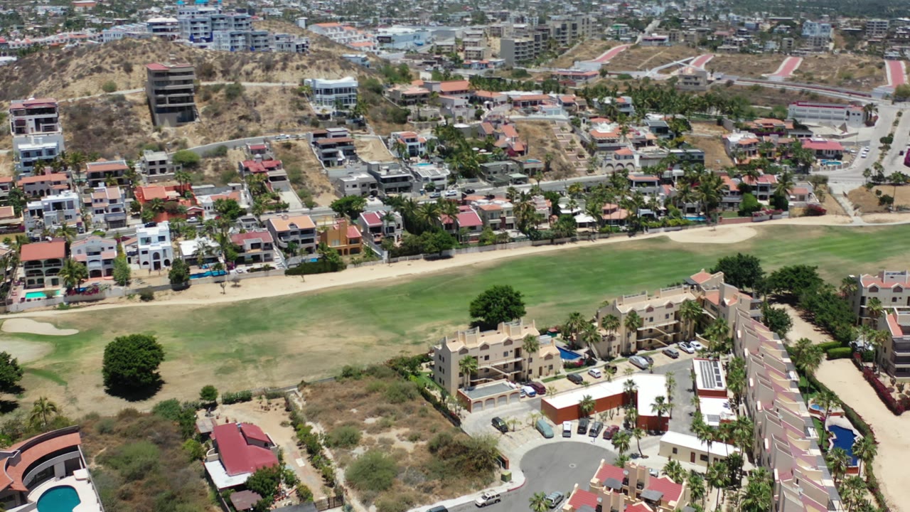 Aerial frame in Los Cabos, Mexico, with a broad green fairway-like area across the lower half, residential buildings and roads in the middle, dry hills in back, and no visible people.