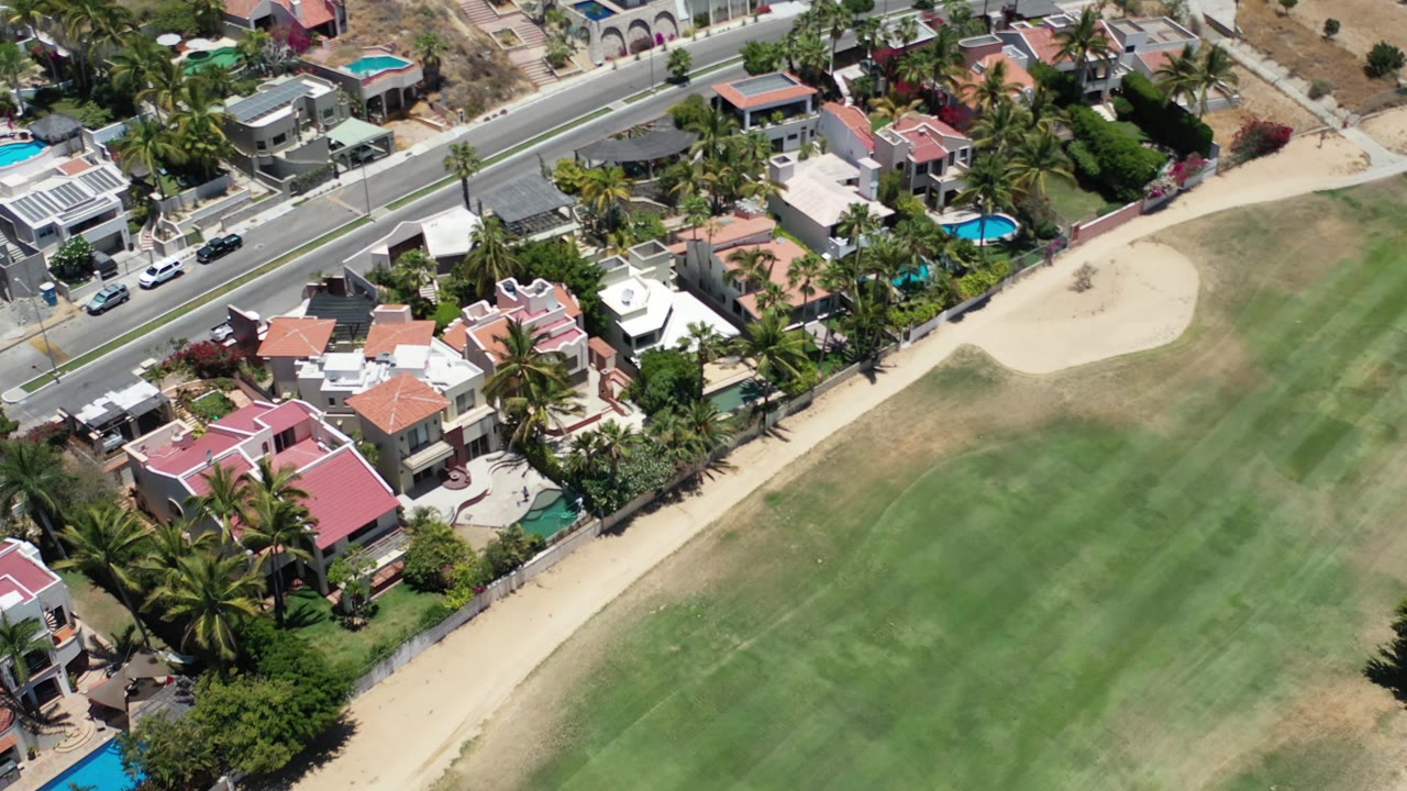 The Los Cabos aerial view shifts slightly over red-roof houses, a roadside line of homes, palms, blue pools, fairway grass, and a sand bunker with no visible people.