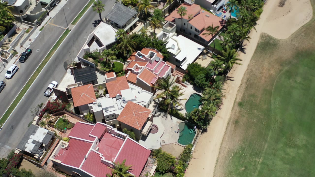 Top-down aerial frame in Los Cabos, Mexico showing red and white roofed homes, palms, a residential road, parked cars, a green landscaped strip, and no visible people.