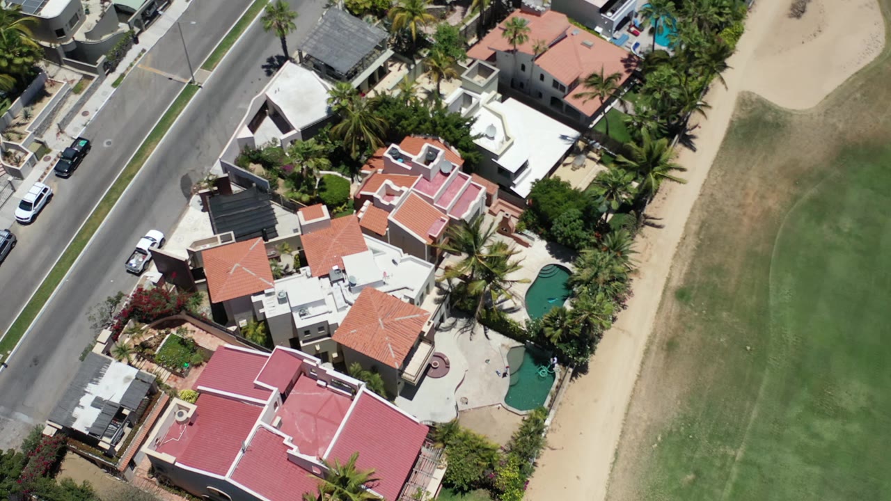 Overhead Los Cabos, Mexico view with the same private homes centered more tightly, a gray road at left, palms around pools, green landscaping at right, and no visible people.
