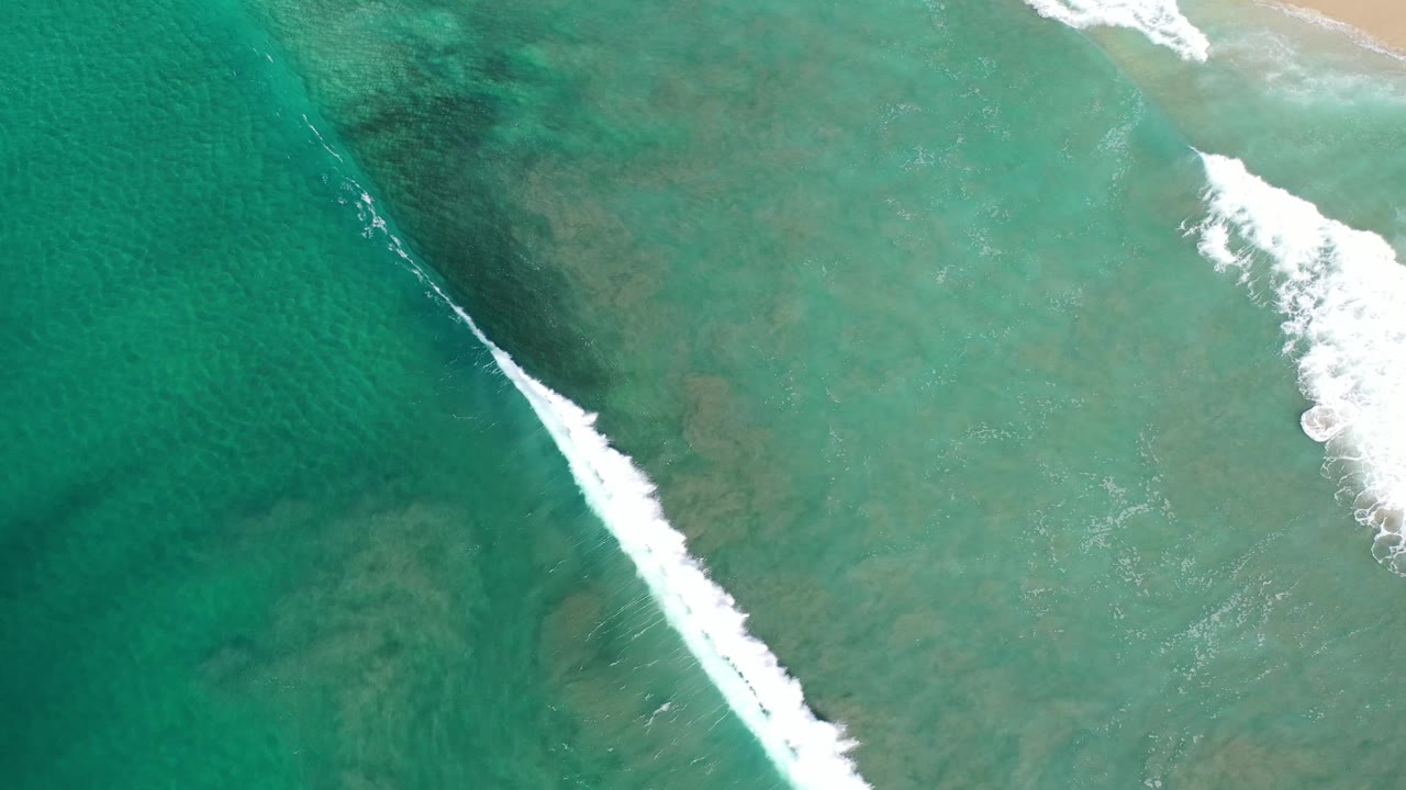 Aerial Los Cabos shoreline frame with green-blue shallow water, a sharper diagonal white surf line, faint sand near the top, and no visible people.