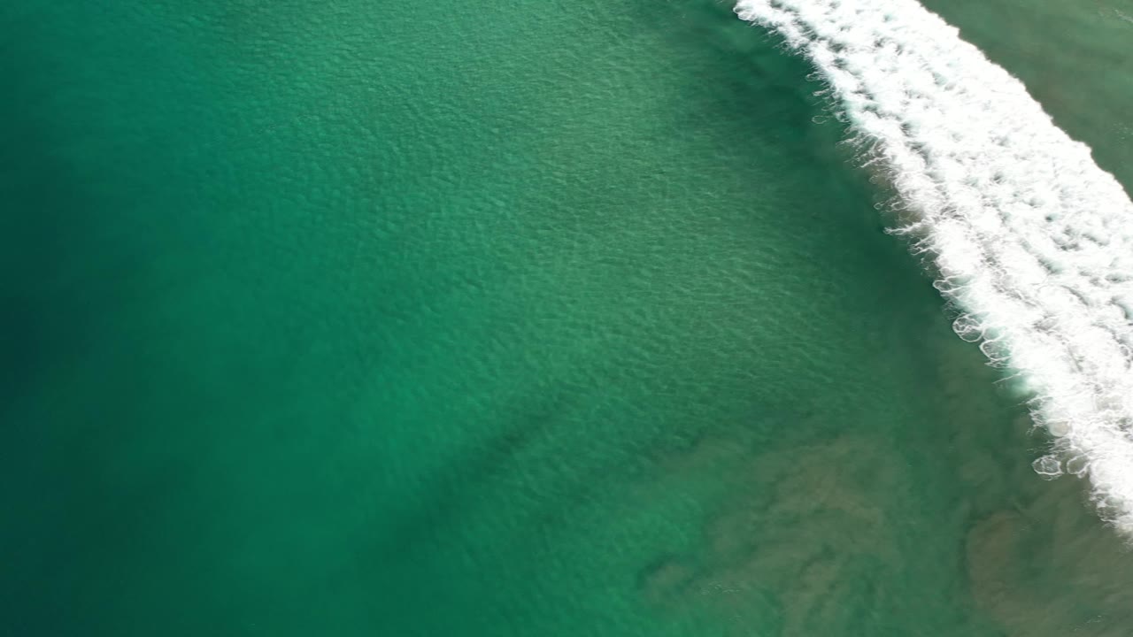 High aerial frame over green-blue ocean in Los Cabos, Mexico, with white surf sliding near the top right edge and no visible people.