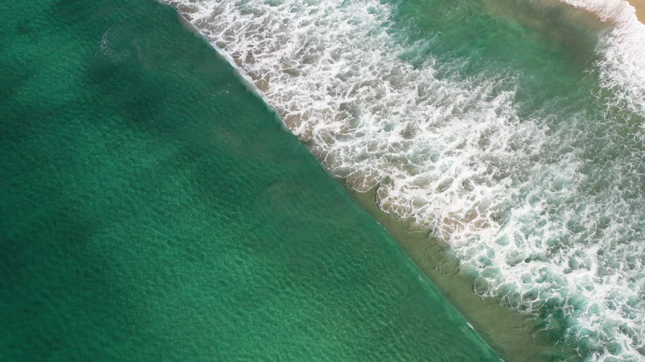 Overhead view of green-blue ocean and diagonal white surf near the Los Cabos shoreline, with no visible people.