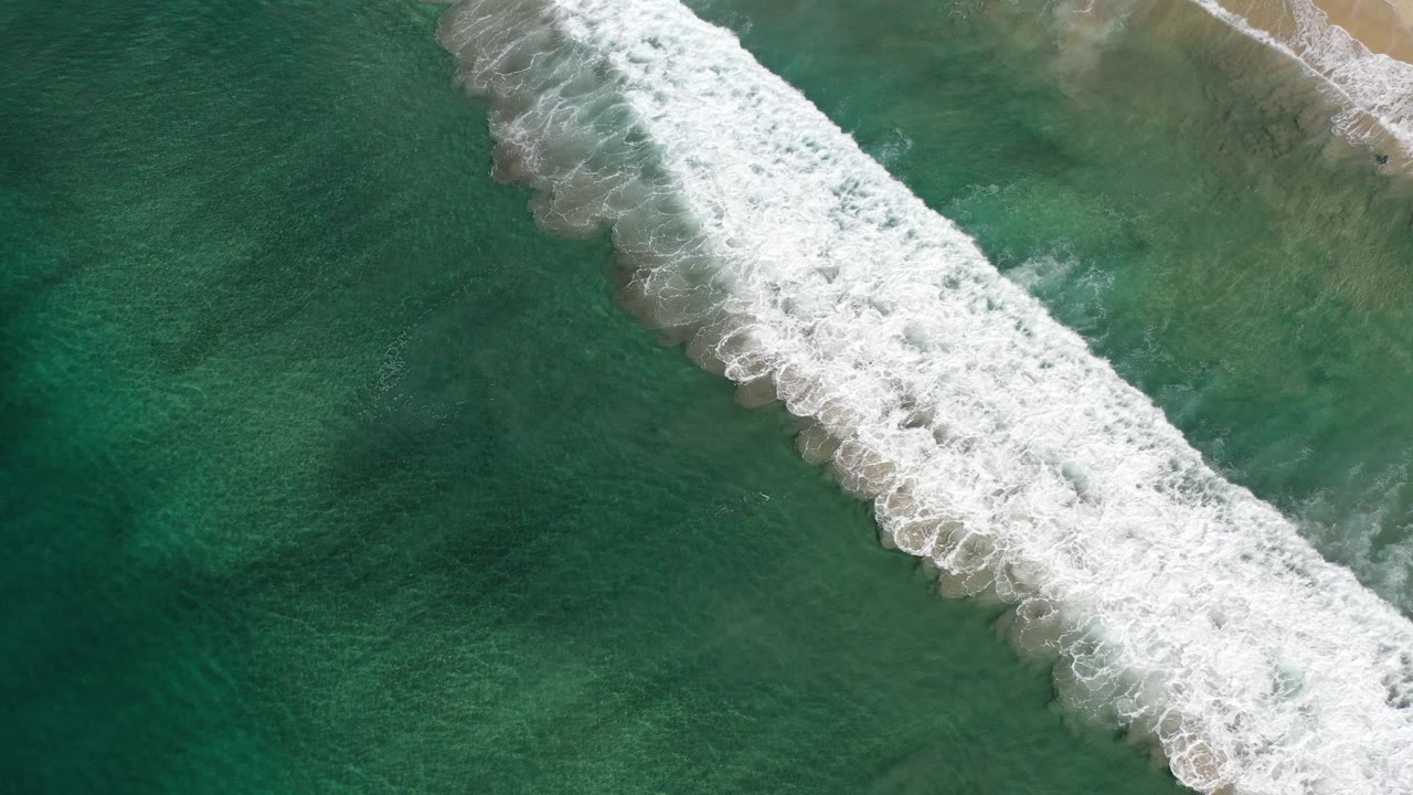 Aerial view over Los Cabos, Mexico, with a diagonal strip of white surf breaking across dark green and teal water, no visible people.