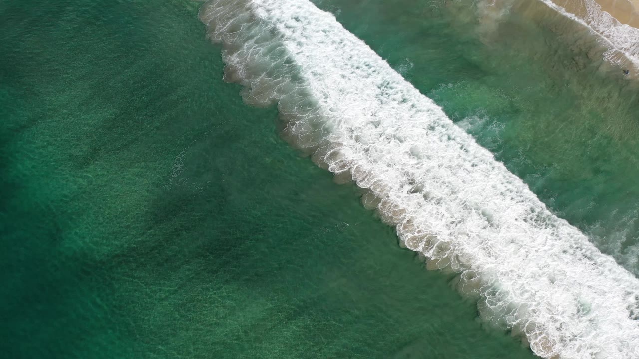 Aerial Los Cabos frame with foamy whitewater stretching diagonally from upper center to lower right over layered green ocean, no visible people.