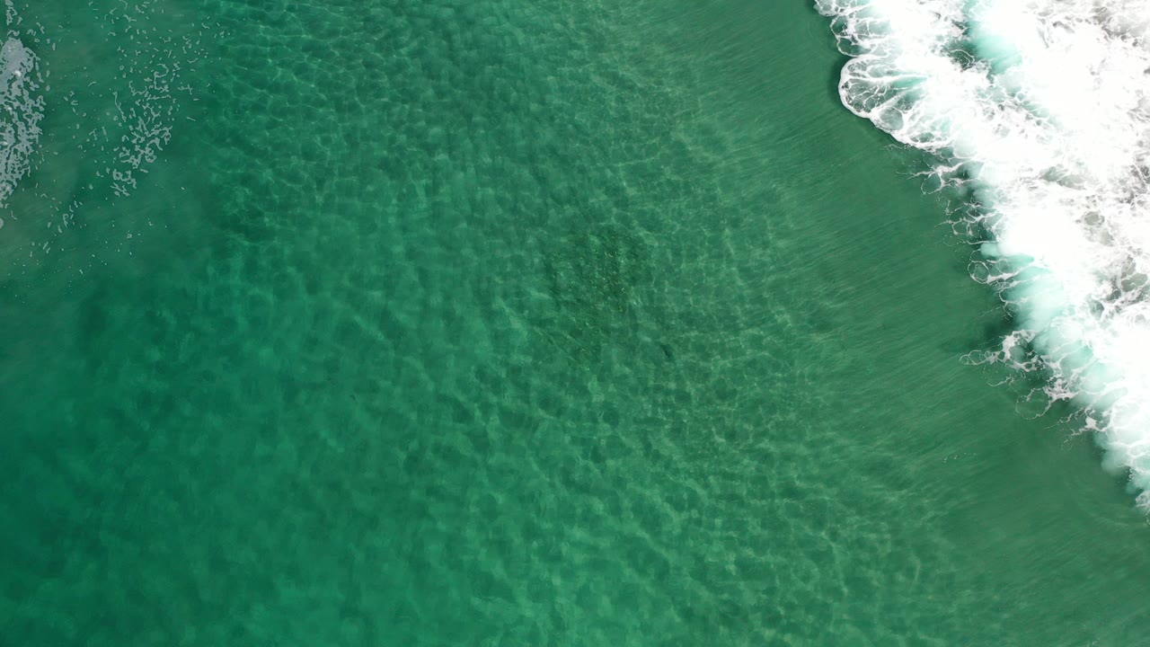 Overhead view of clear turquoise water in Los Cabos, Mexico, with white surf pushing in from the upper right and no visible people.