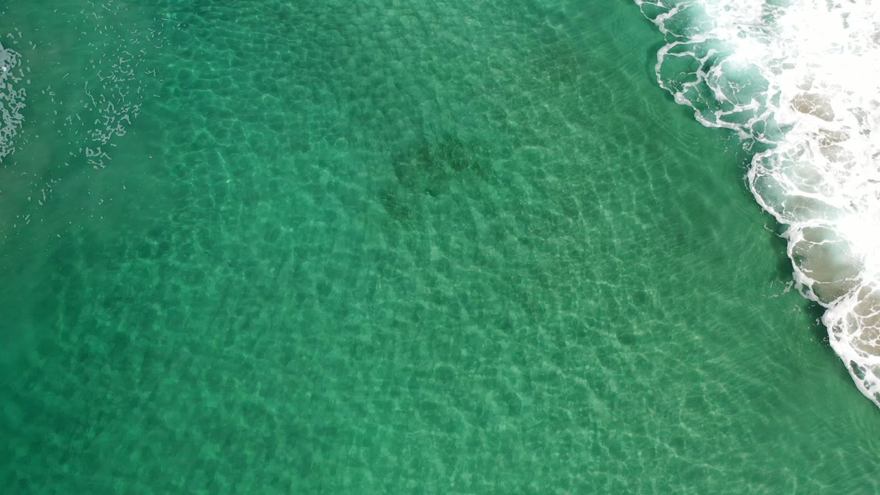 Aerial frame of clear green water in Los Cabos with a narrow band of breaking foam and sand on the right, no visible people.