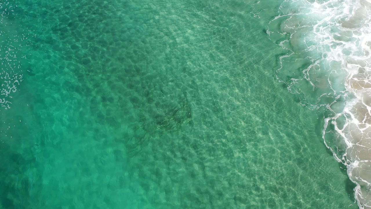Overhead view of clear turquoise water in Los Cabos, Mexico, with faint sand patterns below the surface, white surf entering along the right edge, and no visible people.