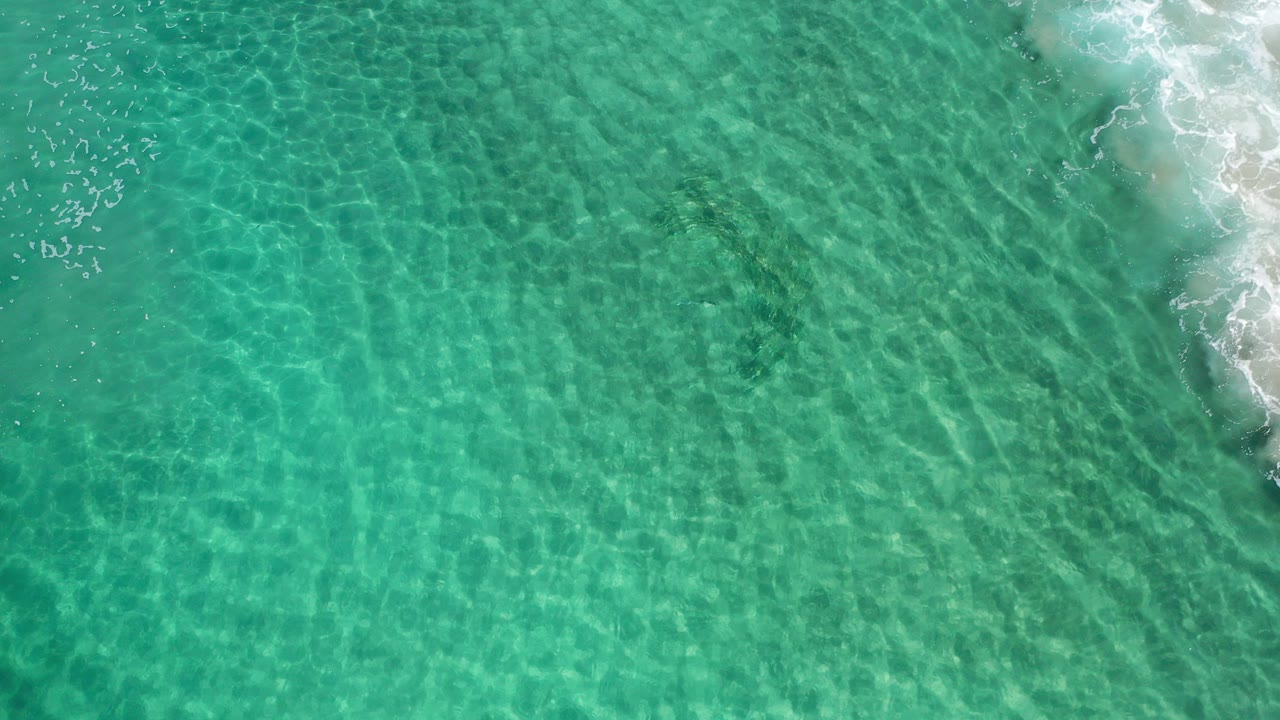 Overhead frame from Los Cabos, Mexico, showing clear teal seawater with bright ripple patterns, faint darker underwater patches, foam along the right edge, and no visible people.