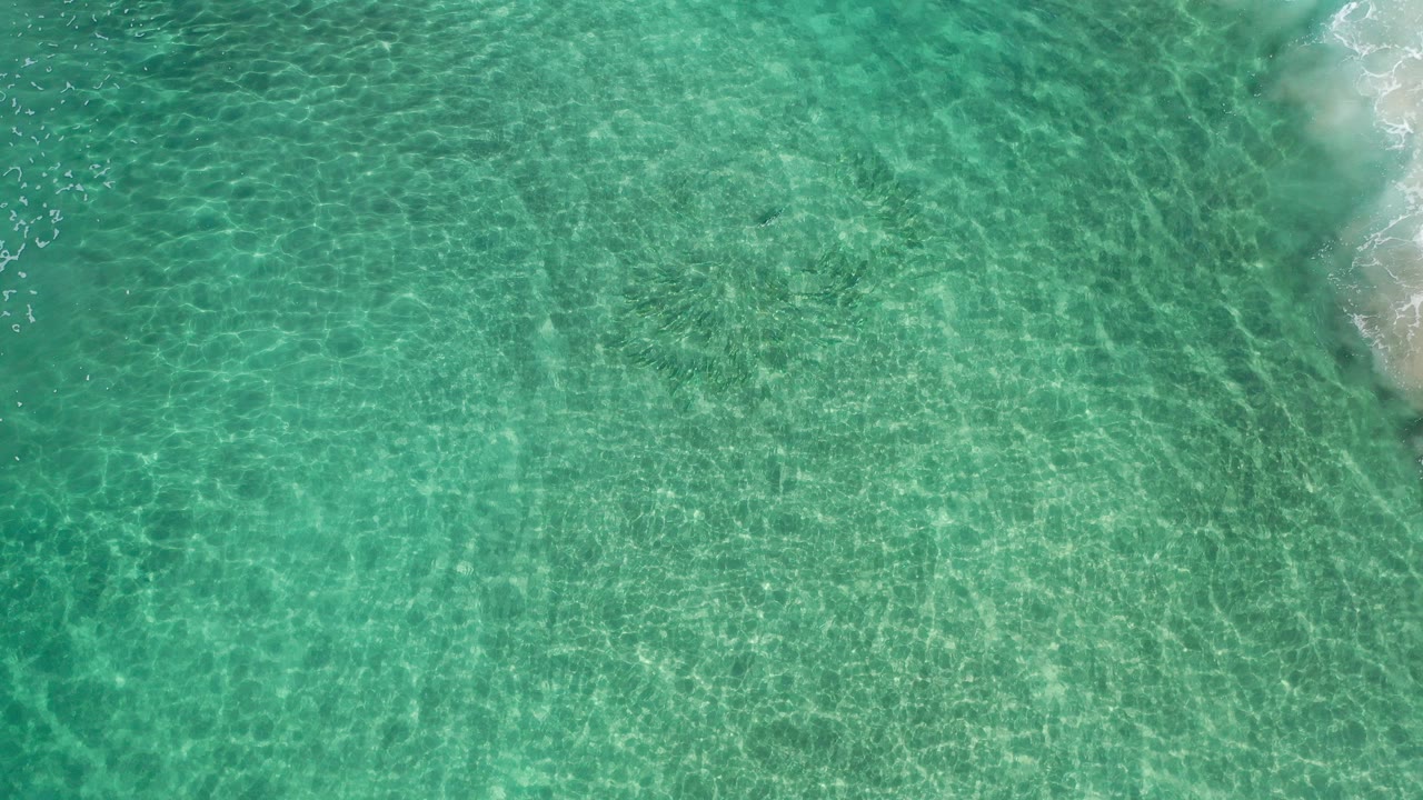 Overhead view of turquoise ocean water in Los Cabos, Mexico, with pale sand and white foam along the upper right edge and no visible people.