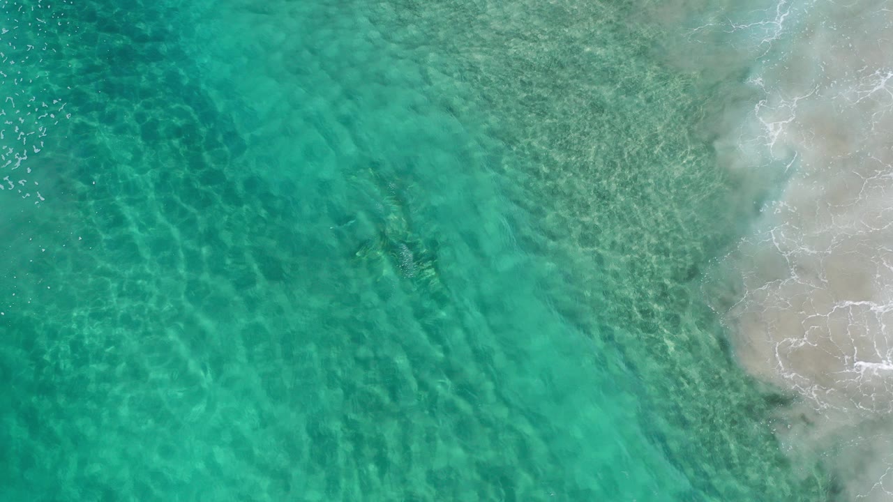 Overhead view of turquoise shallow water meeting pale sand in Los Cabos, Mexico, with faint wave foam at the right edge and no visible people.