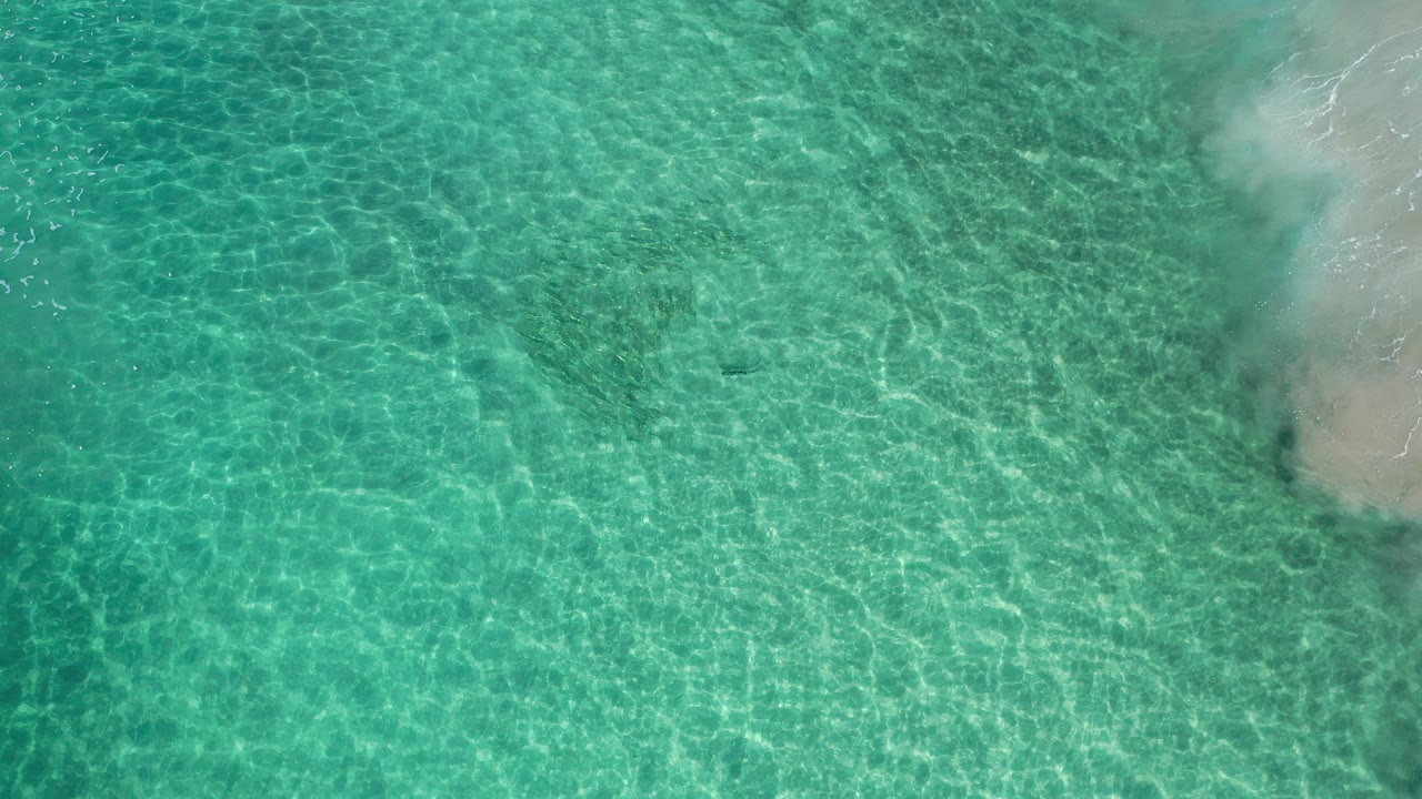 Top-down view of clear turquoise water in Los Cabos, Mexico, with sun ripples across the shallows, a sandy edge at right, and no visible people.