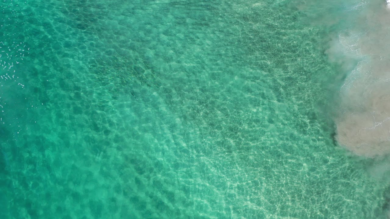 Top-down turquoise water with rippled sand and pale shoreline at right.