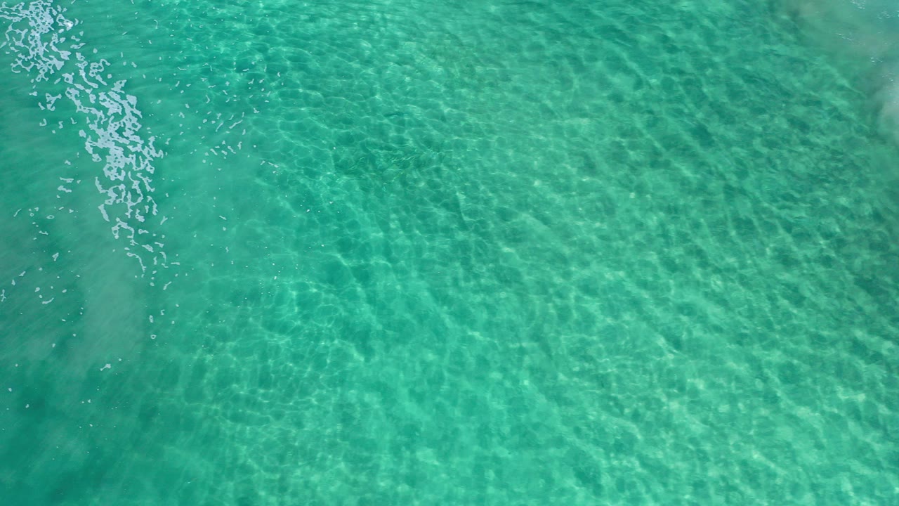 Overhead view of clear turquoise sea water in Los Cabos, Mexico, with rippled sunlight, a darker band near the center, and no visible people.