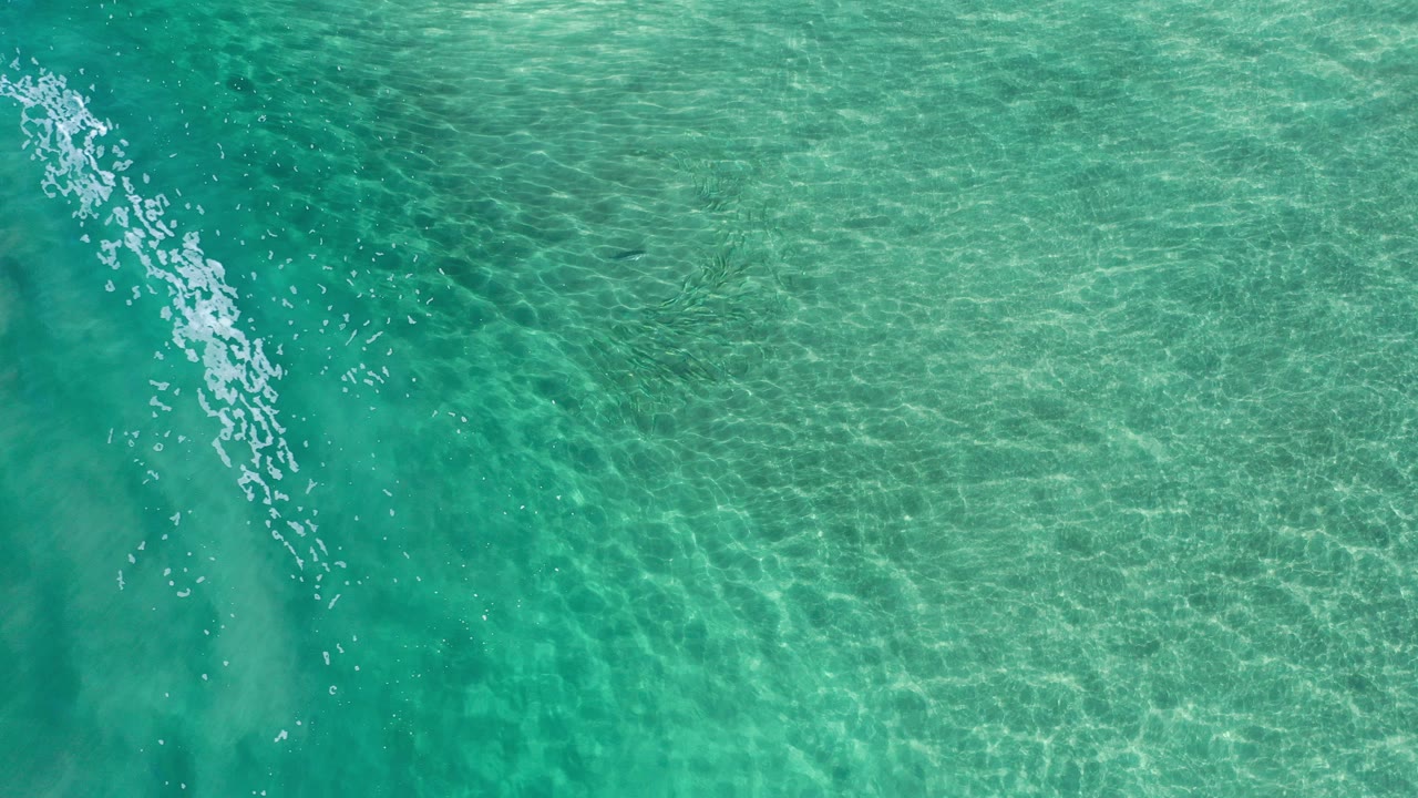 Top-down view of turquoise-green ocean water in Los Cabos, Mexico, with a diagonal band of white foam near the left side and no visible people.