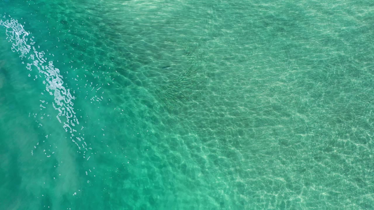 Aerial Los Cabos ocean water fills the frame, with pale foam shifting across the upper-left area and no visible people.