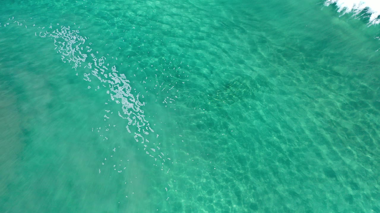 Overhead frame of clear turquoise beach water in Los Cabos with white foam entering near the upper edge, pale sand ripples below, and no visible people.