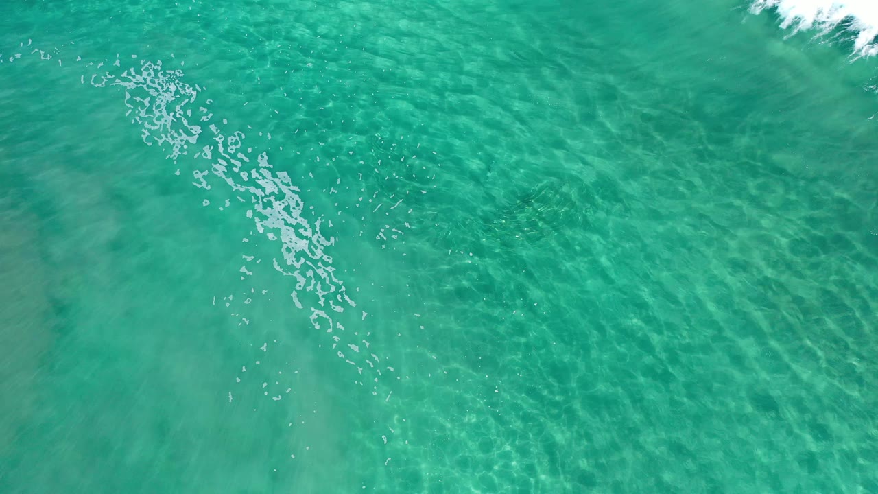 Clear green-blue water in Los Cabos fills the frame, with a diagonal cluster of foam, visible sand ripple lines, and no visible people.