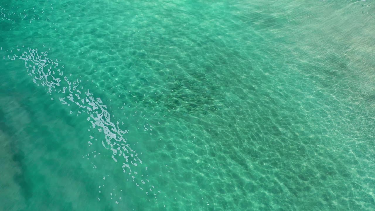 Overhead Los Cabos ocean frame with green-blue water, a longer diagonal reflection pattern, darker submerged areas, and no visible people.