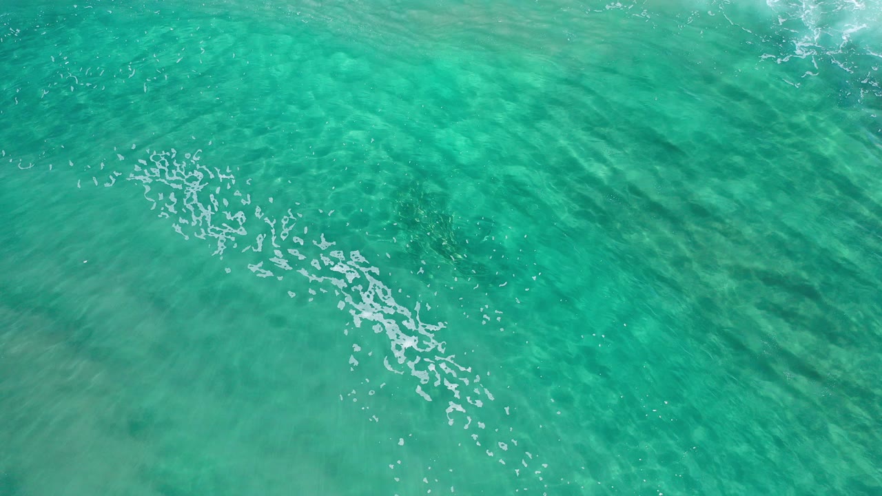 Overhead view of clear turquoise ocean water in Los Cabos, Mexico, with white foam near the left side, rippled sand visible below, and no visible people.