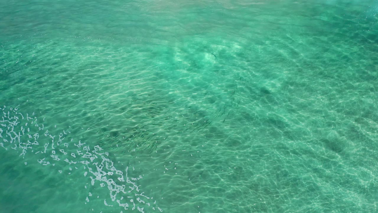 Overhead frame of Los Cabos blue-green water with shimmering surface patterns, soft underwater tonal bands, faint foam marks, and no visible people.