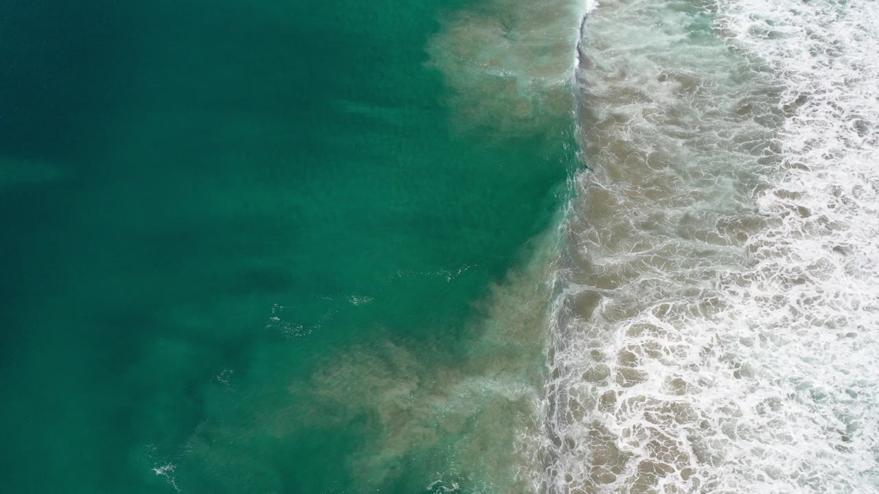 Overhead view in Los Cabos, Mexico showing teal water on the left and white surf breaking over pale sand on the right, with no visible people.