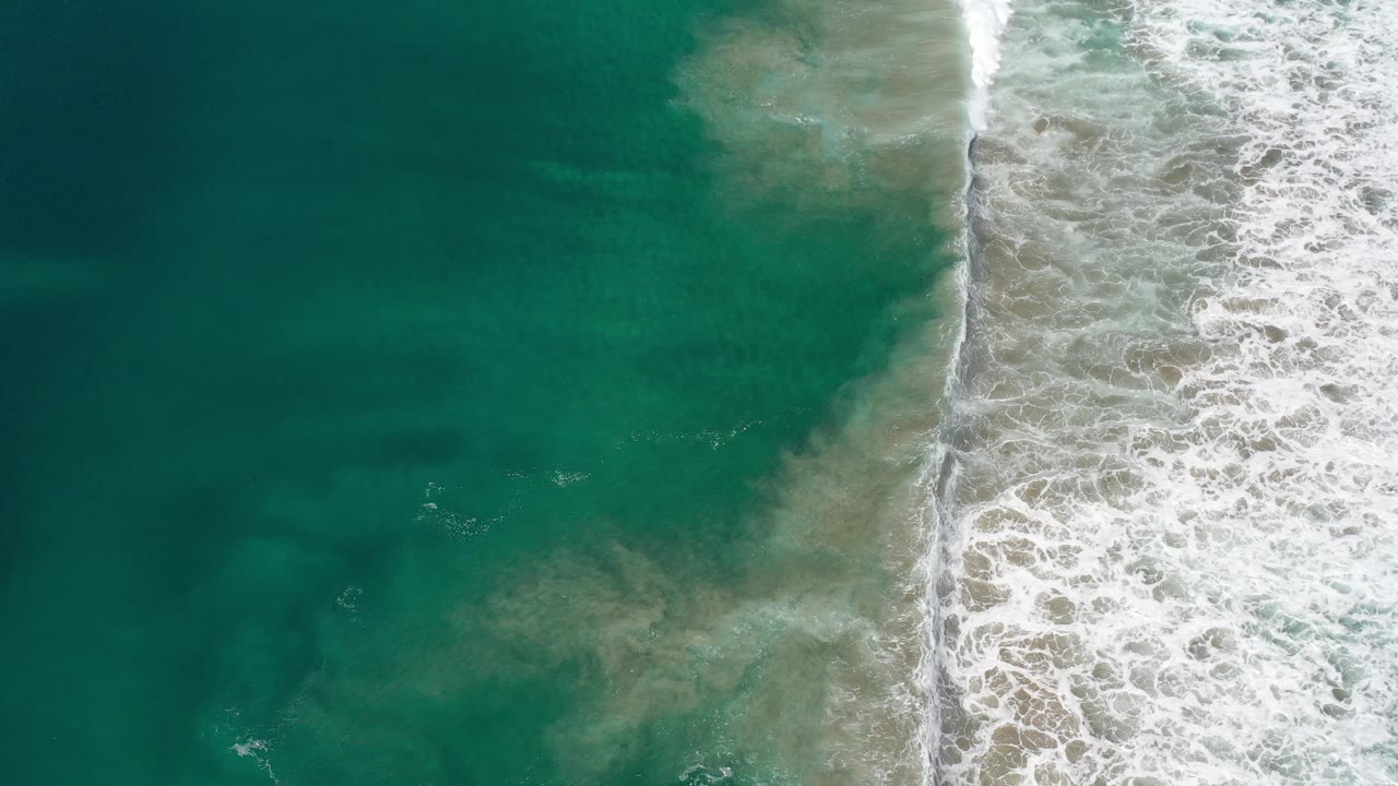 White surf curls over sand next to open teal water.