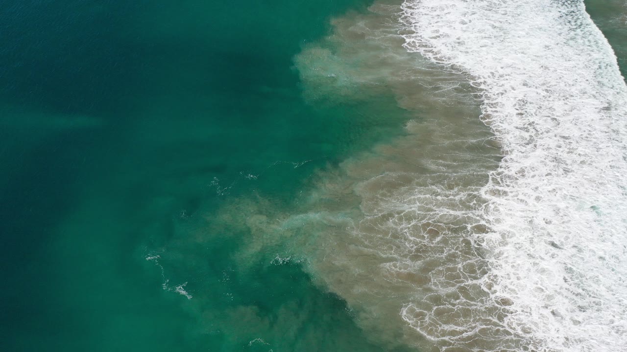 Overhead view in Los Cabos, Mexico, with teal water on the left and white surf spreading over sand on the right, no visible people.