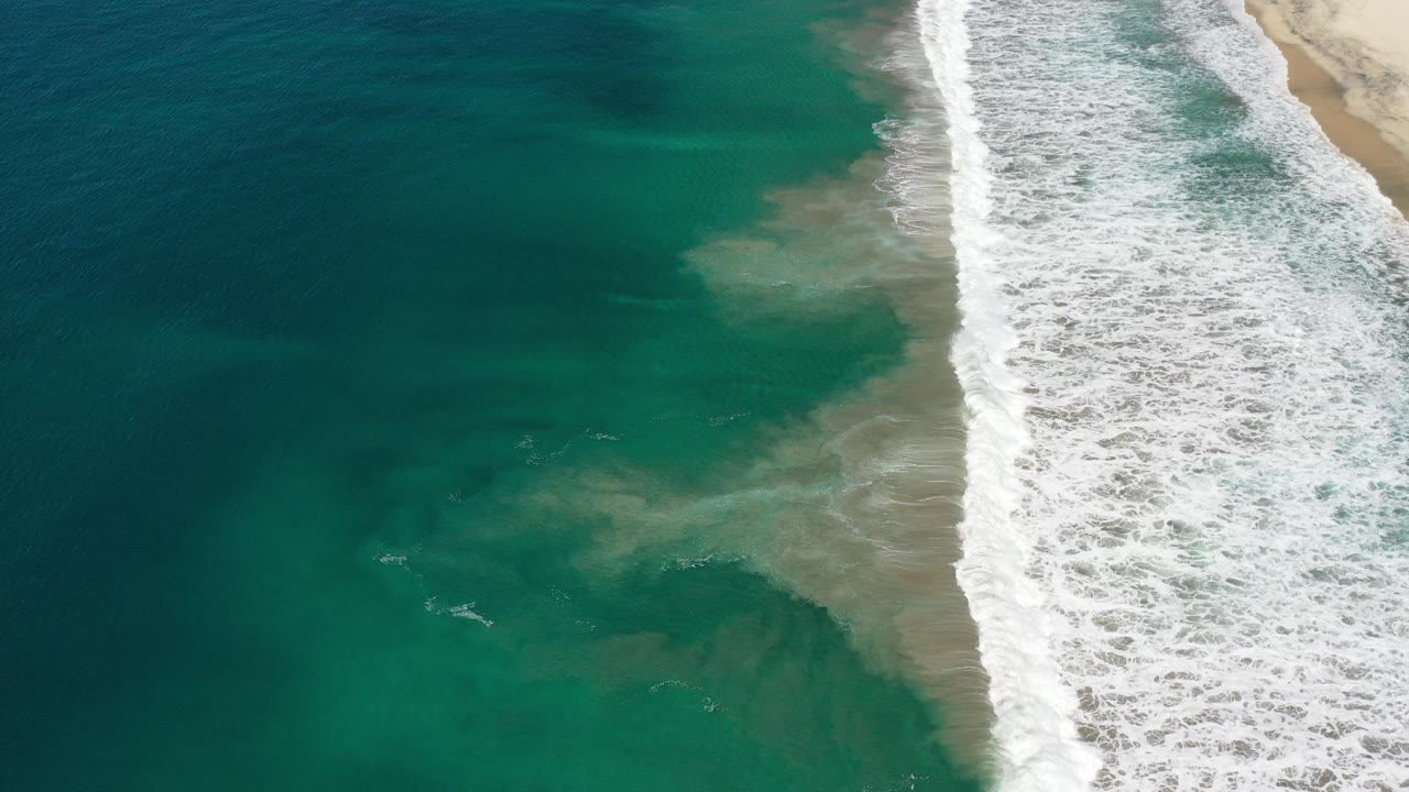 Aerial view over the Los Cabos coast with turquoise water, a diagonal strip of white surf, pale beach at the right edge, and no visible people.