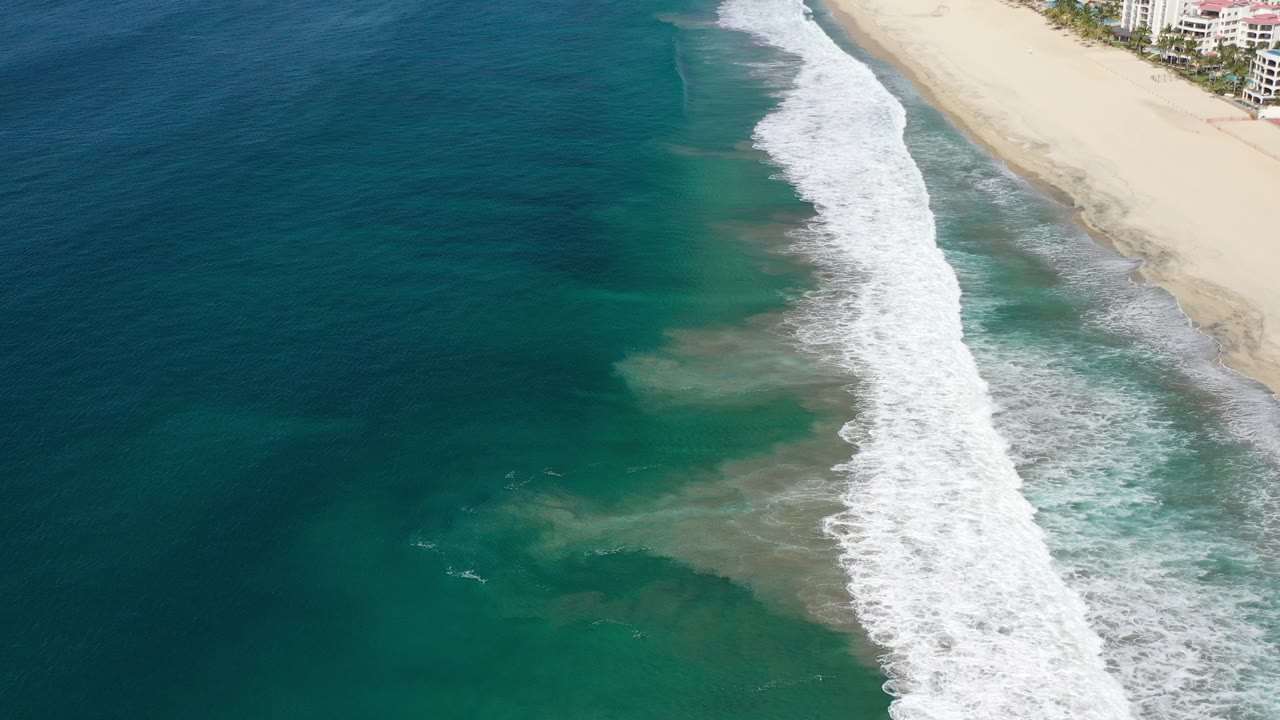 Aerial view of teal ocean waves breaking beside a sandy beach and coastal buildings.